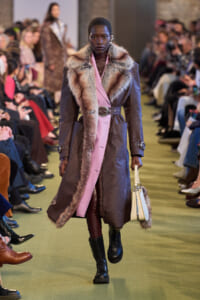 Model walking the runway in a brown fur-trimmed coat with pink lining, belt at the waist, and a light handbag in hand.