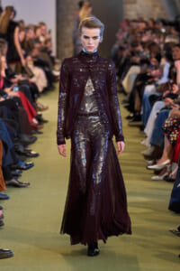Model walks a fashion runway in a shiny burgundy sequined suit with a sheer, flowing cape-like overlay.