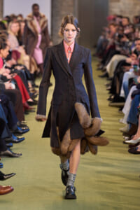 Model walks the runway in a dark, double-breasted wool coat with fur trim at the cuffs and hem, pink shirt beneath, and metallic shoes. Audience lines the catwalk take photos.