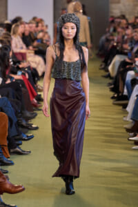 Model walks a fashion show wearing a sparkly beaded hat, a glittery camisole top, and a high-waisted burgundy leather-like skirt down the runway.