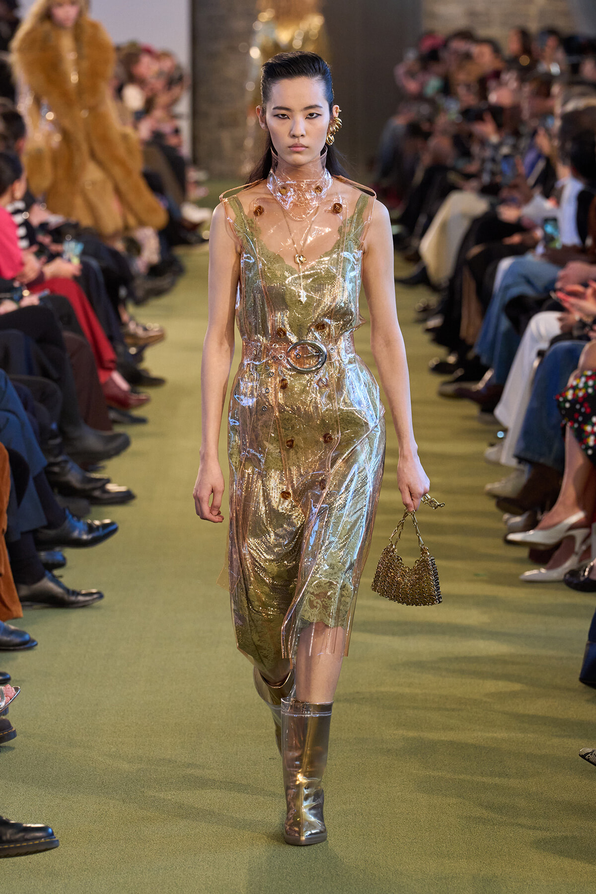 Model on a runway wearing a metallic, translucent green-silver dress with a wide belt, knee-high metallic boots, and a small beaded handbag.