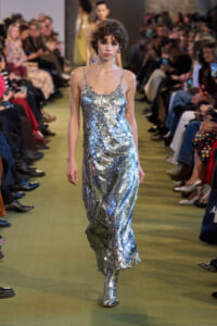 Model walks a runway in a silver-sequined slip dress, audience on both sides at a fashion show caped by green floor.