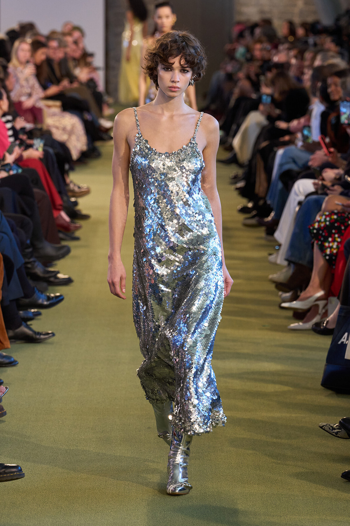 Model walks a runway in a silver-sequined slip dress, audience on both sides at a fashion show caped by green floor.