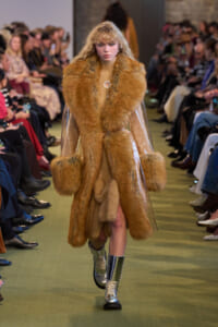 Model walking the runway in a long tan fur coat with oversized fur cuffs and metallic boots.