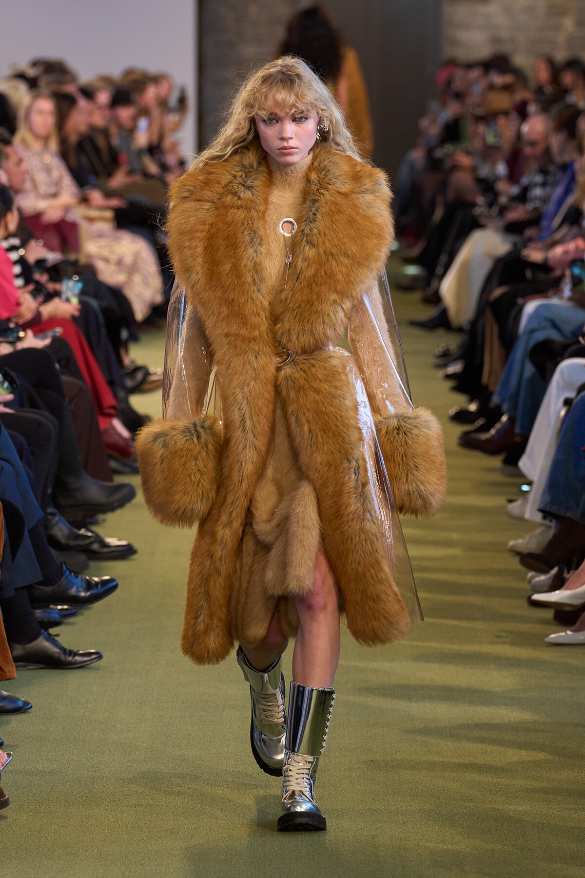 Model walking the runway in a long tan fur coat with oversized fur cuffs and metallic boots.