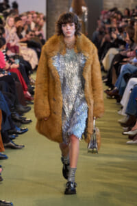 Model walking the runway in a brown fur coat over a shimmering silver dress, holding a small metallic handbag.