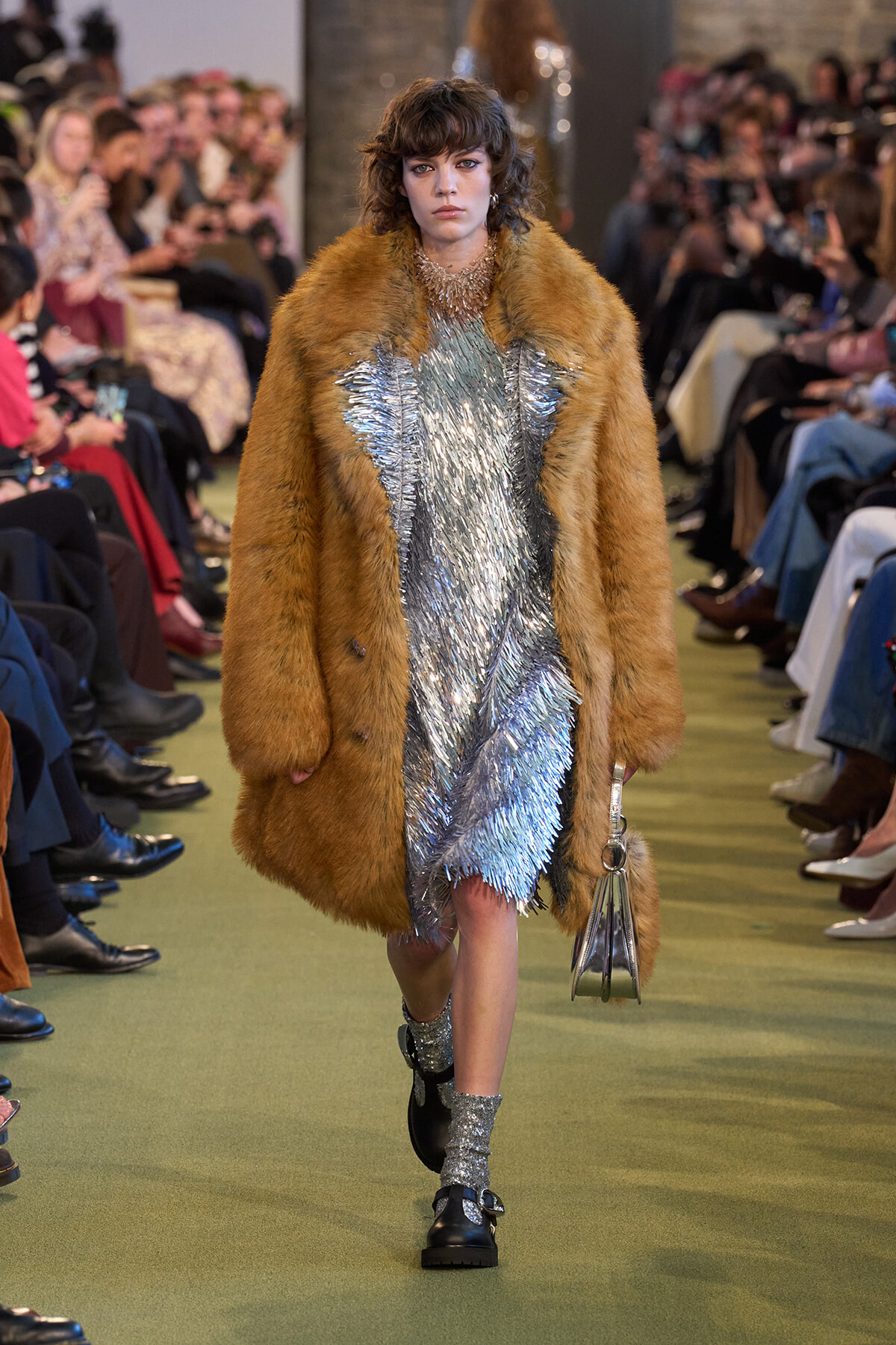 Model walking the runway in a brown fur coat over a shimmering silver dress, holding a small metallic handbag.