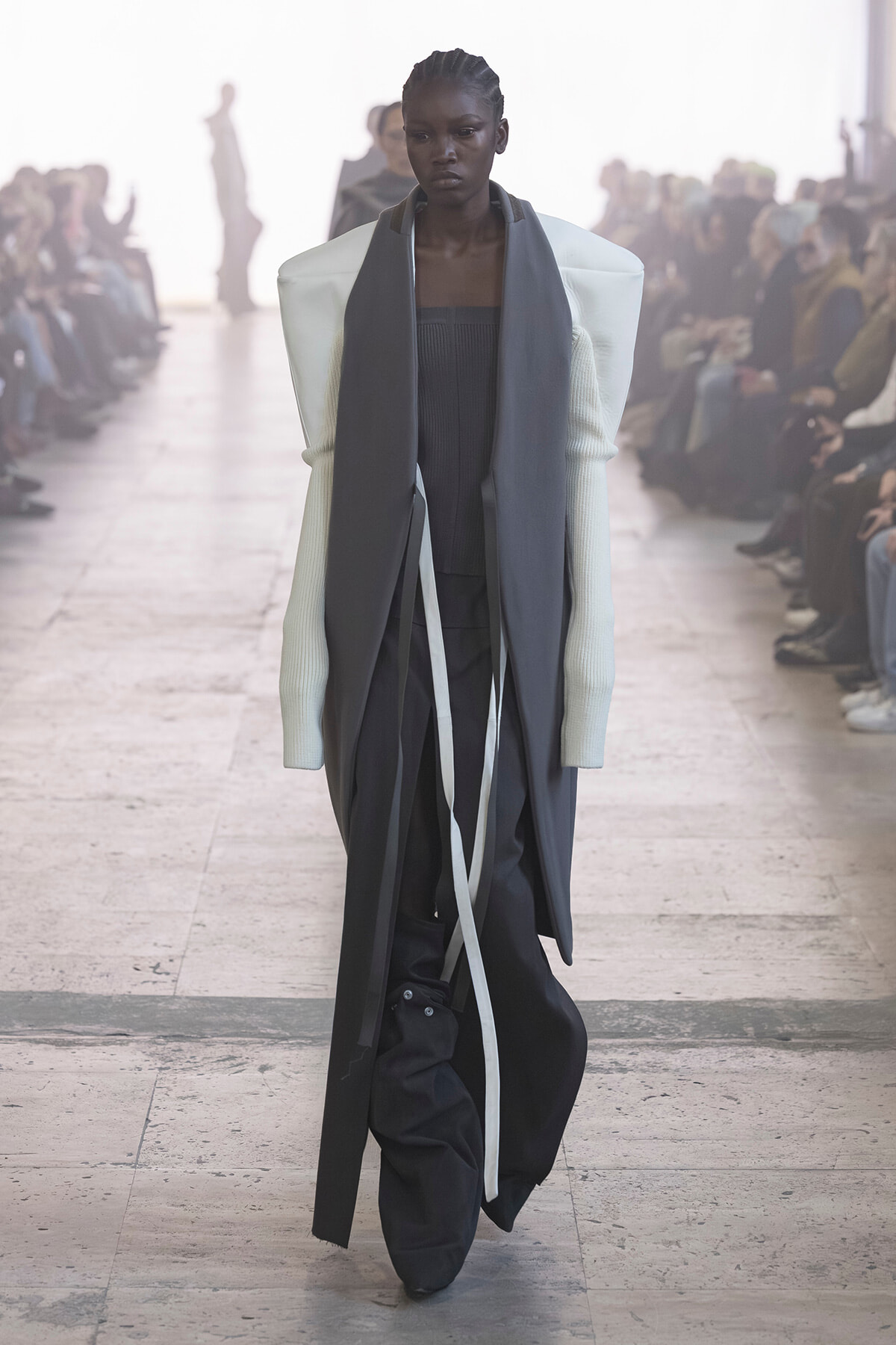 Model walks a fashion runway in an avant-garde layered outfit: white-sleeved top with dark gray draped panels and long straps over black pants.