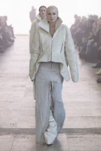 Model walks a runway in a white sequined oversized jacket with a high collar and zip pockets, paired with textured wide-leg pants, audience in the background.