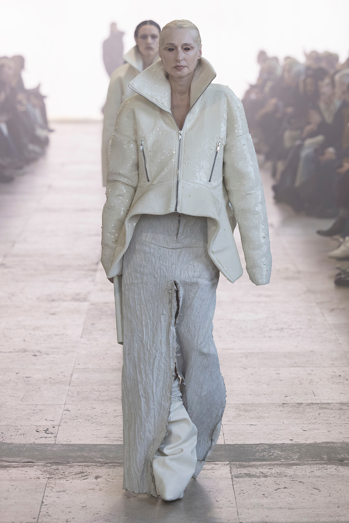 Model walks a runway in a white sequined oversized jacket with a high collar and zip pockets, paired with textured wide-leg pants, audience in the background.