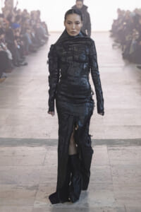 Model walks a runway wearing a textured, all-black, high-neck gown with distressed, frayed detailing and a front slit. Audience blurred in background.
