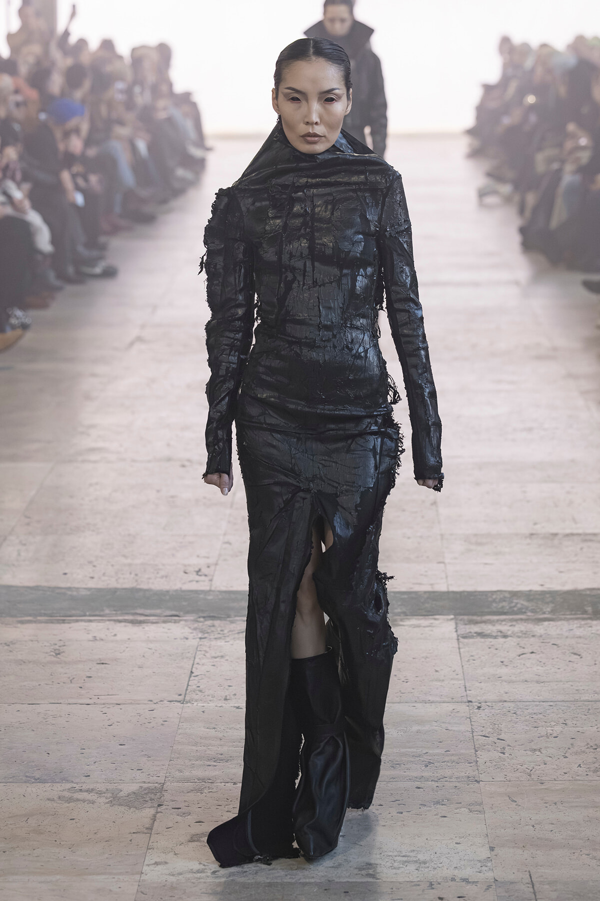 Model walks a runway wearing a textured, all-black, high-neck gown with distressed, frayed detailing and a front slit. Audience blurred in background.