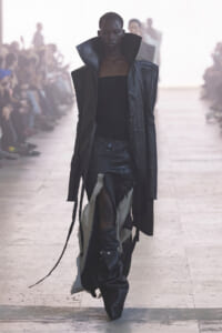 Model walks a fashion runway in a dark, oversized leather coat with high collar and torn black pants. Audience blurred in background.