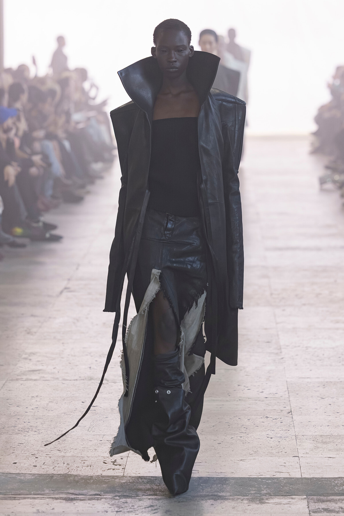 Model walks a fashion runway in a dark, oversized leather coat with high collar and torn black pants. Audience blurred in background.