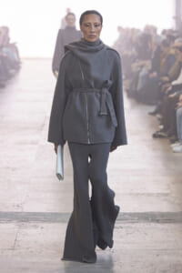 Model walking a fashion show in a charcoal gray wrap coat with belt, matching knit pants, and a light clutch; audience blurred in the background.