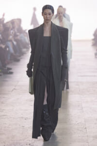 Model walks the runway in a dark, layered ensemble with broad shoulders and long scarf, audience blurred in the background.