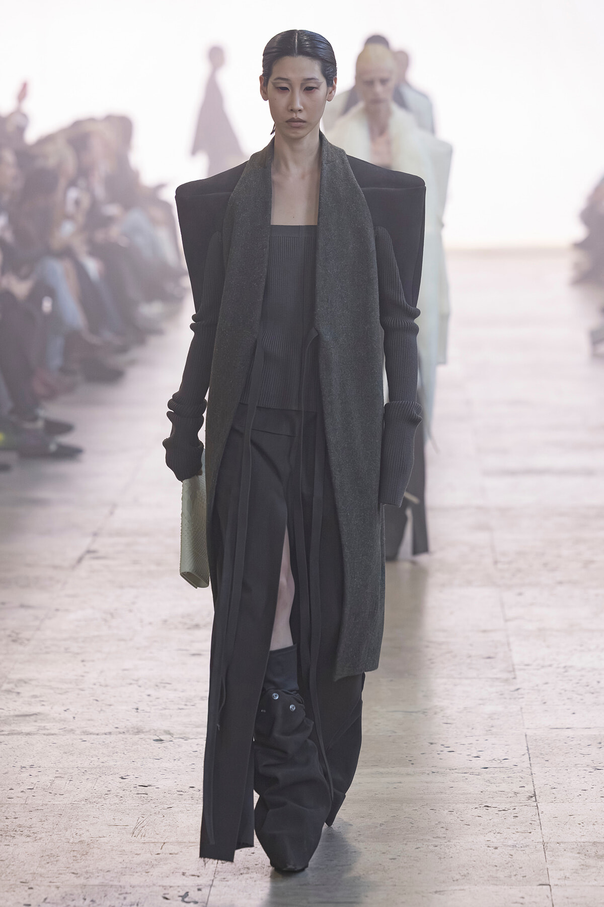 Model walks the runway in a dark, layered ensemble with broad shoulders and long scarf, audience blurred in the background.