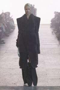 Model on a runway wearing a long black shearling vest over a structured black outfit with exaggerated shoulders.