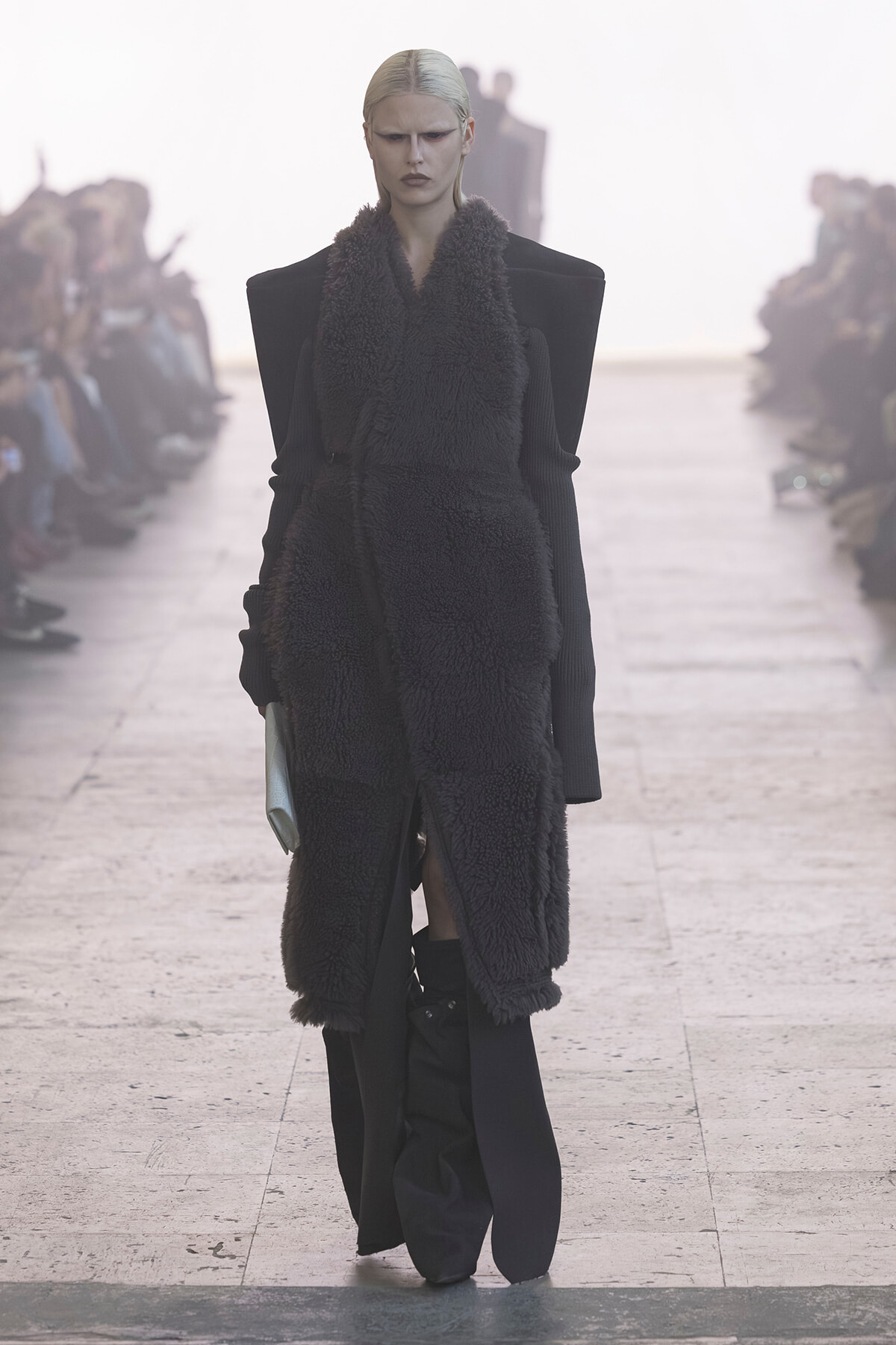 Model on a runway wearing a long black shearling vest over a structured black outfit with exaggerated shoulders.