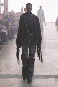 Model walks a fashion runway wearing a black fringe cape-like garment with a belt, paired with textured fur-like pants and long fringe detailing at the sleeves and legs.