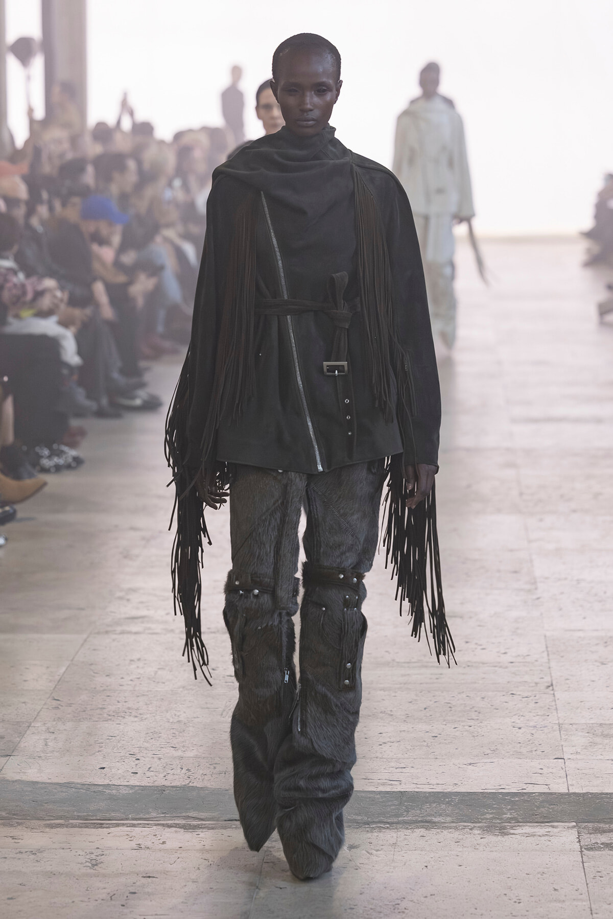 Model walks a fashion runway wearing a black fringe cape-like garment with a belt, paired with textured fur-like pants and long fringe detailing at the sleeves and legs.