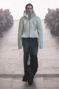 Model walking a runway in a pale mint oversized jacket with a high collar and distressed blue jeans on the catwalk.
