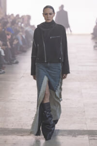 Model walking a runway in a black zippered jacket, layered over a distressed denim skirt with a long, frayed hem and tall black boots.