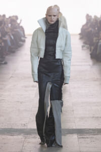 Bald model walks a runway in a light quilted cropped jacket over a black textured outfit; audience blurred in the background.