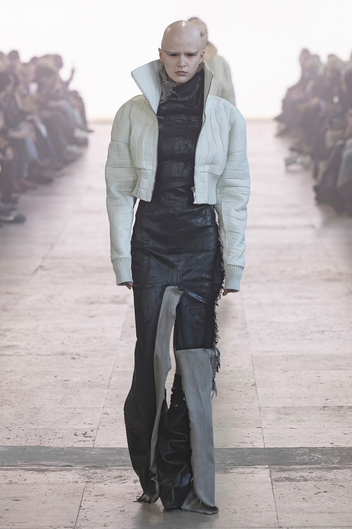 Bald model walks a runway in a light quilted cropped jacket over a black textured outfit; audience blurred in the background.