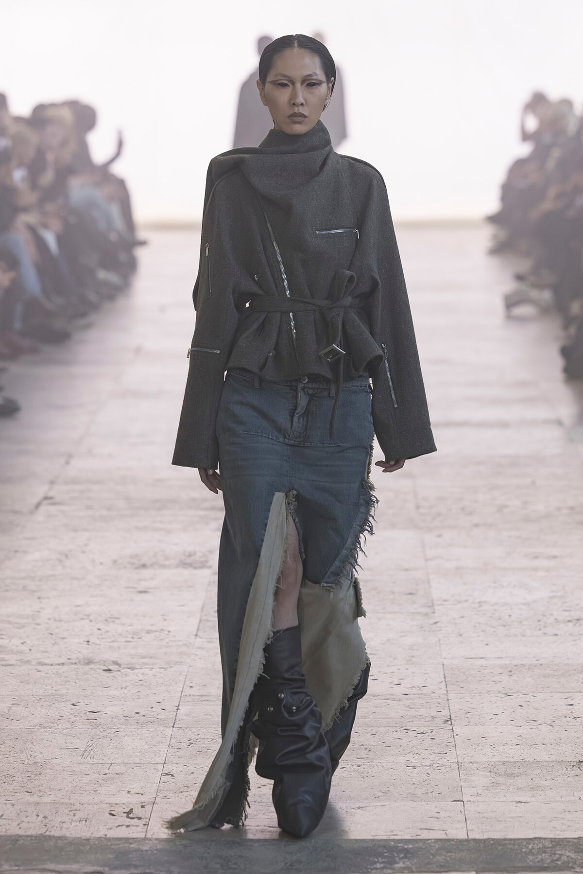 Model walks down a runway in a dark textured coat with a belt, paired with ripped jeans and tall boots, during a fashion show.