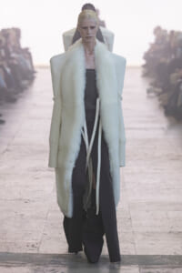 Model walks the runway in a long white fur-trimmed coat over a dark outfit, spotlighted on a fashion show catwalk