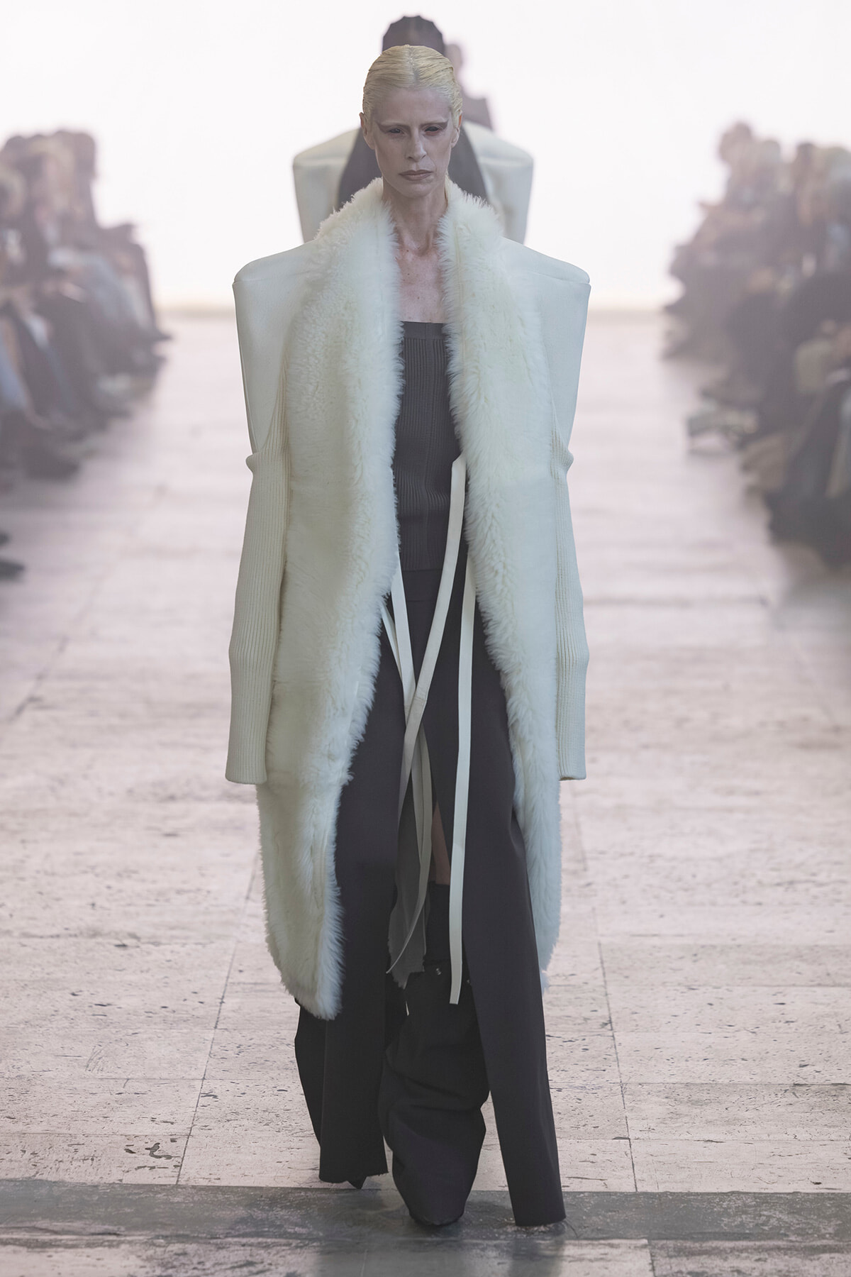 Model walks the runway in a long white fur-trimmed coat over a dark outfit, spotlighted on a fashion show catwalk