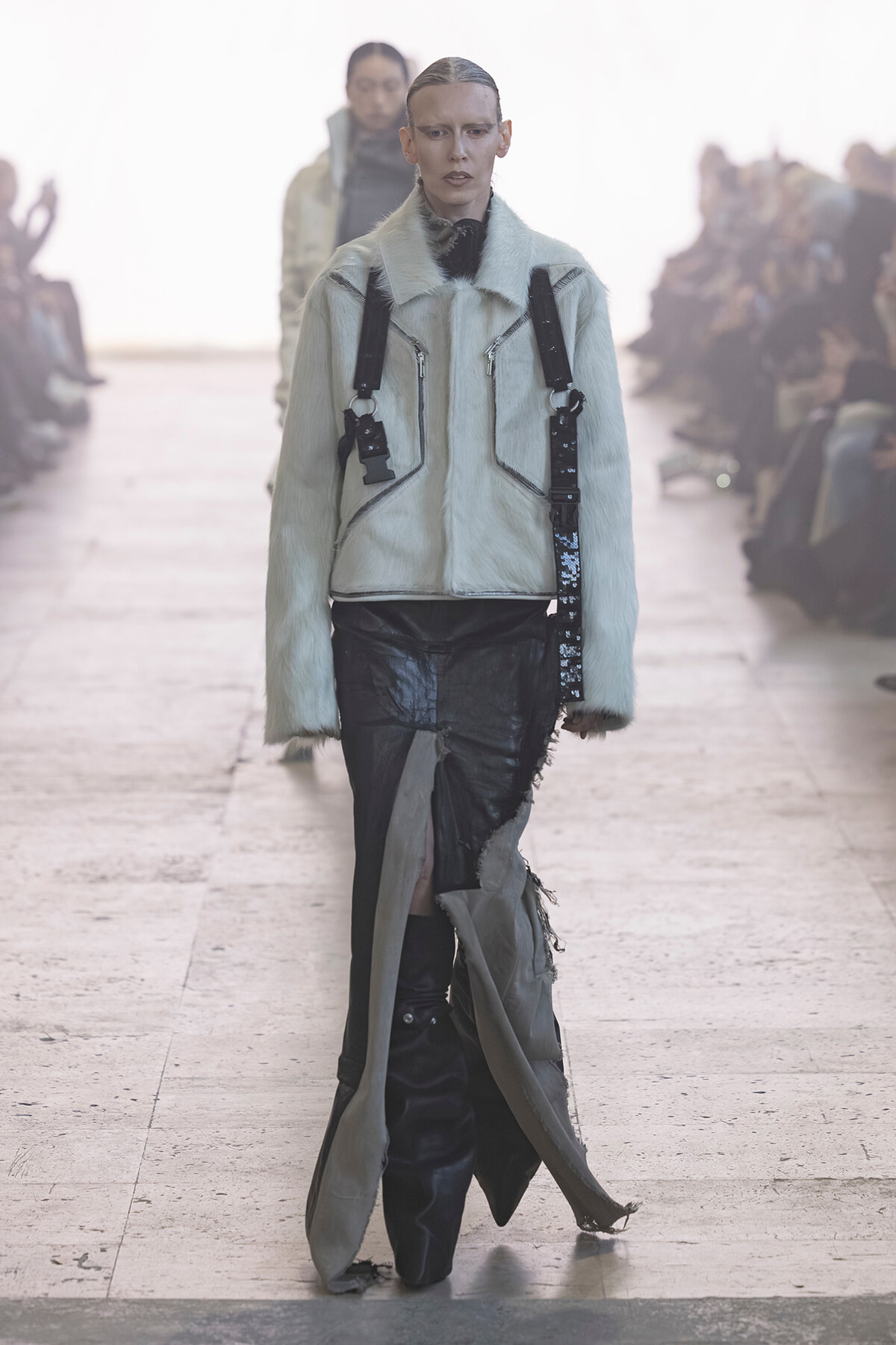 Model walks the runway in a pale blue fur jacket with black utility straps and distressed black pants.
