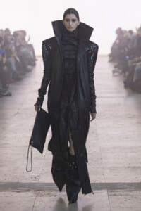 Runway model in an all-black avant-garde outfit with a tall oversized collar, sequined shoulders, and a long coat walking the catwalk, clutch in hand.