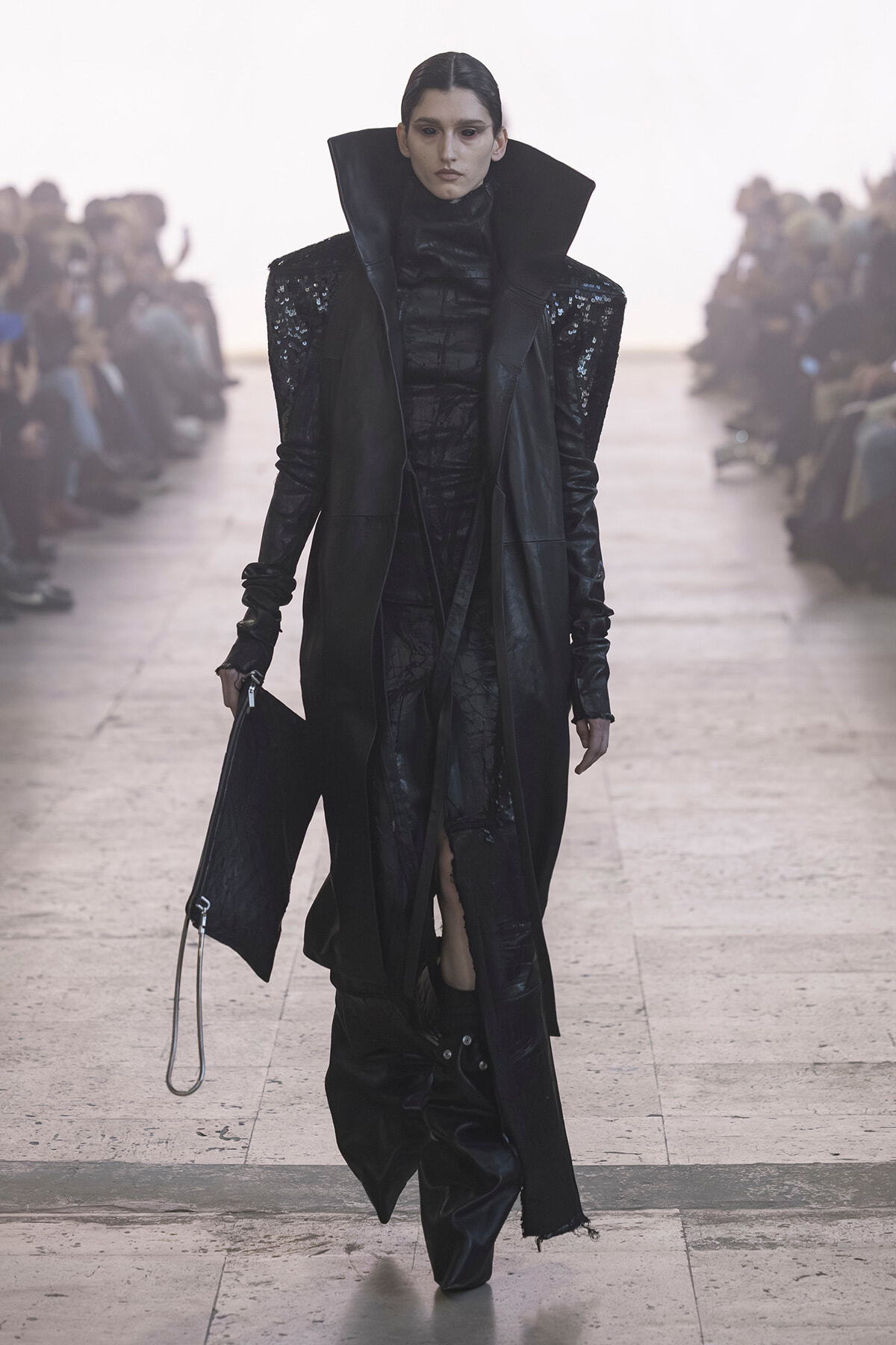 Runway model in an all-black avant-garde outfit with a tall oversized collar, sequined shoulders, and a long coat walking the catwalk, clutch in hand.
