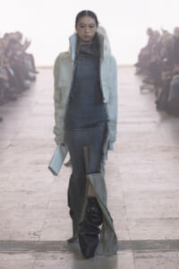 Model walks runway in a pale fur jacket over a denim dress with a long, layered skirt and oversized black boots, holding a white clutch.