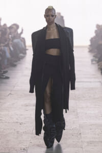 Model on a fashion runway wearing a black oversized coat, bandeau top, high-slit pants, and knee-high boots.