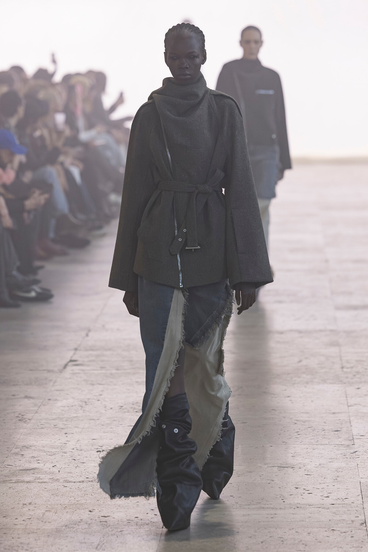 Model walks a runway in a dark, oversized belted coat with a draped, patchwork skirt trailing behind; audience blur in the background.