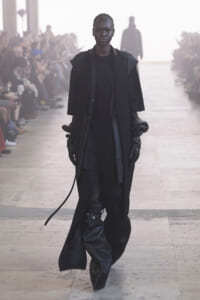 Model walking a runway in an all-black, oversized outfit with long gloves and flowing coat.