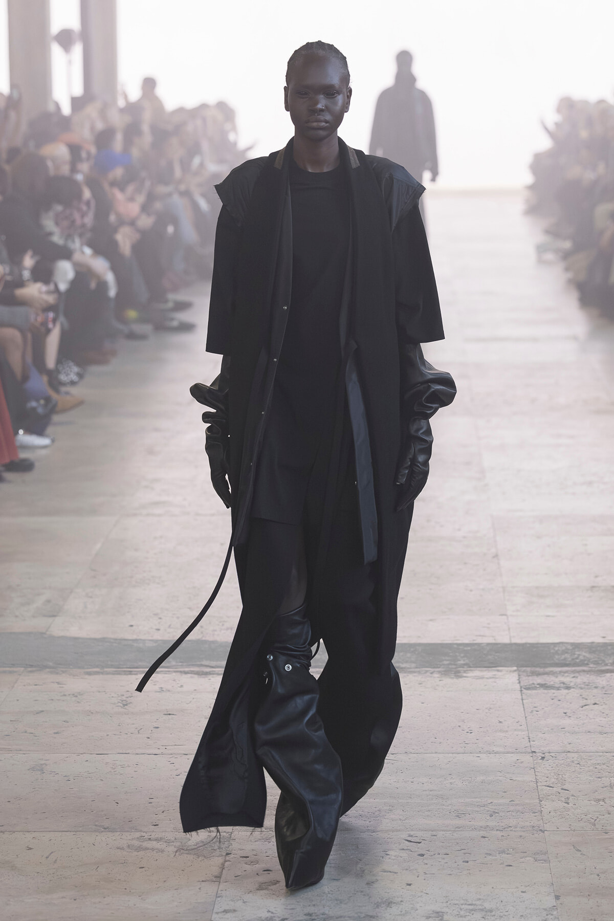 Model walking a runway in an all-black, oversized outfit with long gloves and flowing coat.
