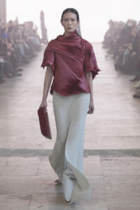 Model walks a runway in a burgundy pleated top and pale gray wide-leg pants, holding a red clutch as spectators watch.