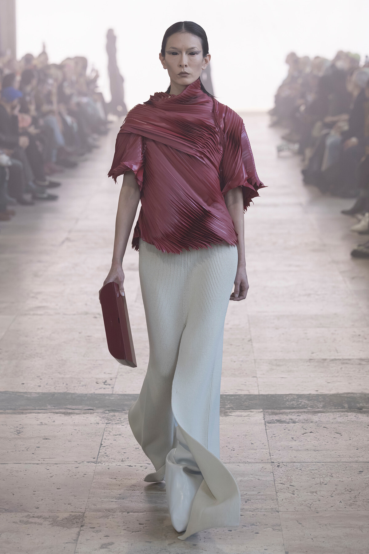 Model walks a runway in a burgundy pleated top and pale gray wide-leg pants, holding a red clutch as spectators watch.