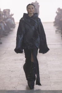 Model walks the runway in a glossy black, oversized pleated coat with a high collar and dramatic sleeves.