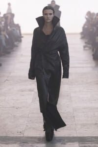 Model walking the runway in a black leather trench with a high collar, dramatic silhouette.