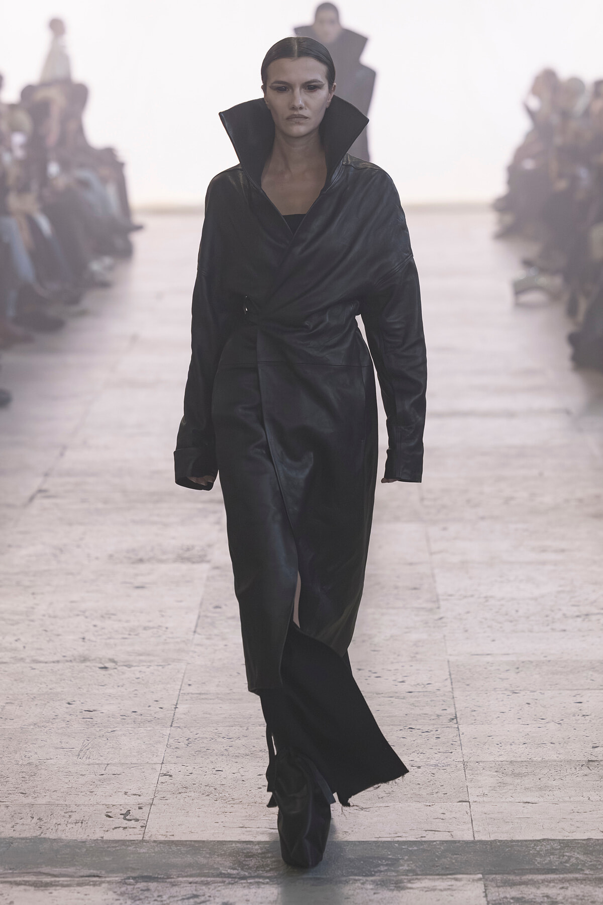 Model walking the runway in a black leather trench with a high collar, dramatic silhouette.