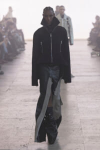 Model walks a runway wearing a black oversized zip-front jacket with a tall stand-up collar and two vertical zippers, paired with distressed layered pants; audience blurred in the background.