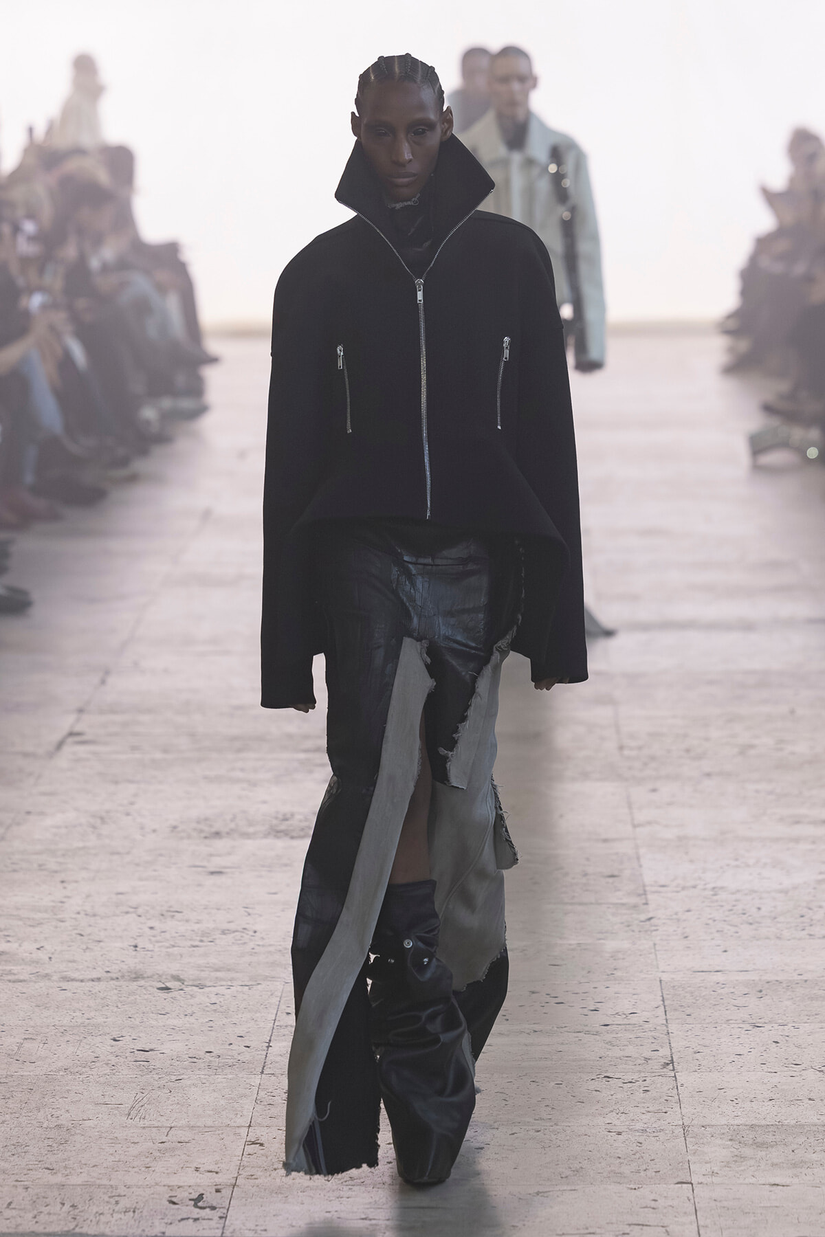 Model walks a runway wearing a black oversized zip-front jacket with a tall stand-up collar and two vertical zippers, paired with distressed layered pants; audience blurred in the background.