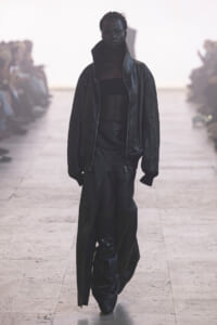 Model walks down a runway in all-black outfit: oversized leather jacket, cropped top, and wide, draped leather pants.