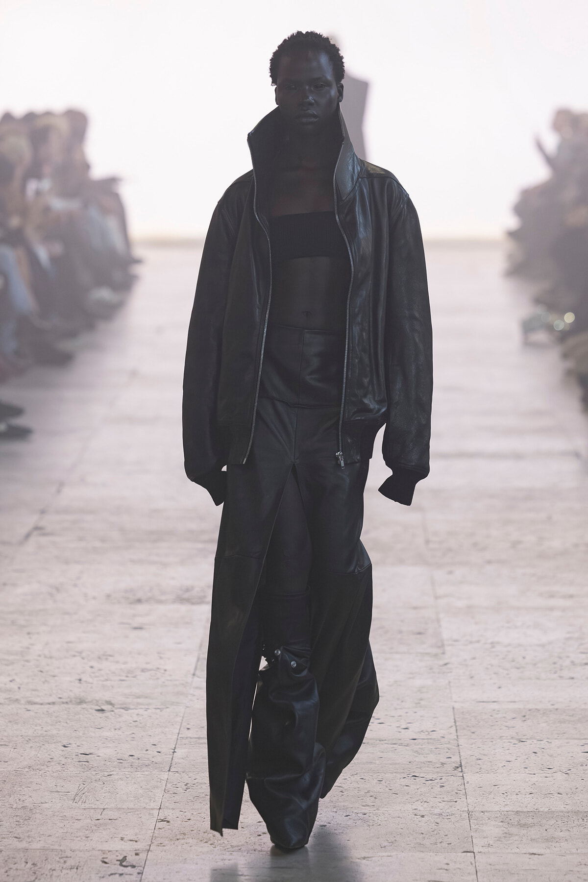 Model walks down a runway in all-black outfit: oversized leather jacket, cropped top, and wide, draped leather pants.