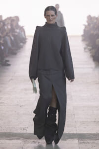 Runway model in a dark gray structured top with oversized sleeves and a front-slit skirt, black boots, and a pale green clutch in hand.